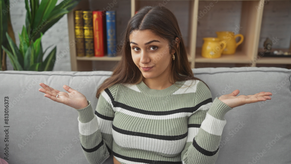 Young hispanic woman with neutral expression, sitting on a sofa indoors, gesturing uncertainty with her hands in a cozy room.