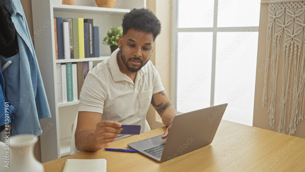 African american man using credit card for online shopping at home on laptop in a well-organized living room. African american man using credit card for online shopping at home on laptop in a well-organized living room.
