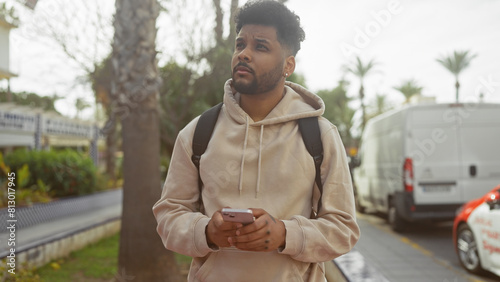 A thoughtful african american man in casual attire with a backpack standing on a city street holding a smartphone.