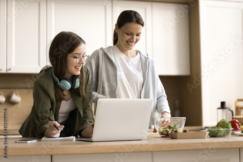 Happy smart teen school girl and positive young mom studying online and cooking salad for dinner at kitchen table, using laptop, chopping vegetables, talking, laughing, having fun