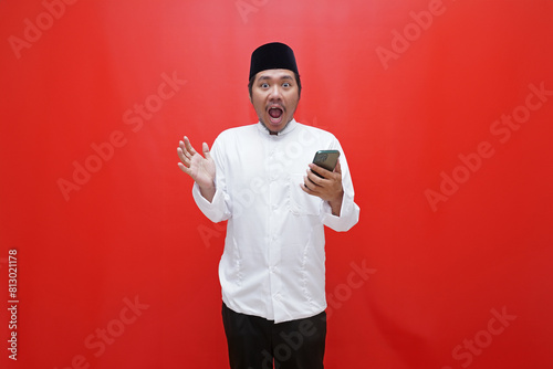 Asian muslim man with surprised expression looking at camera and holding smartphone isolated on red background photo