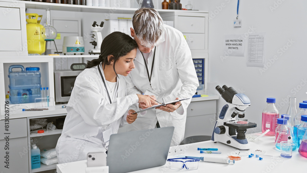 A man and woman, possibly coworkers, intently review data on a clipboard in a well-equipped laboratory setting. A man and woman, possibly coworkers, intently review data on a clipboard in a well-equipped laboratory setting.