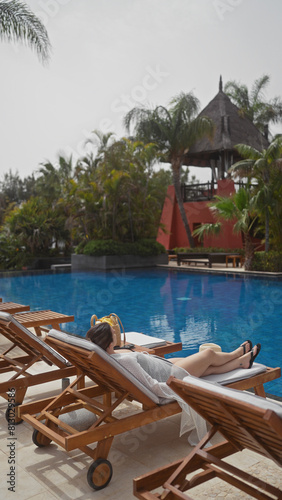 A young woman relaxes on a lounger by a resort s blue pool in bali  surrounded by tropical foliage.
