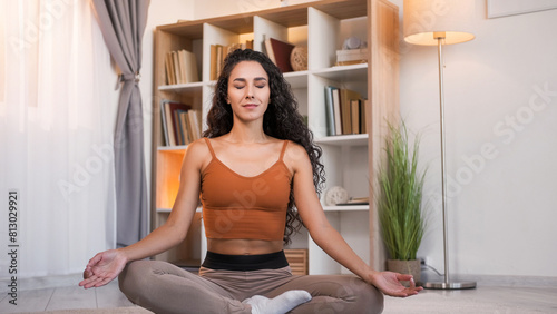 Meditation practice. Yoga relaxation. Peaceful smiling girl enjoying calming exercise sitting cross-legged in lotus pose on floor at light home interior with copy space.