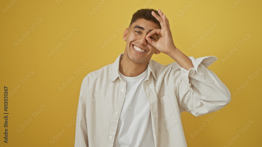 Handsome man making okay gesture over eye against yellow background