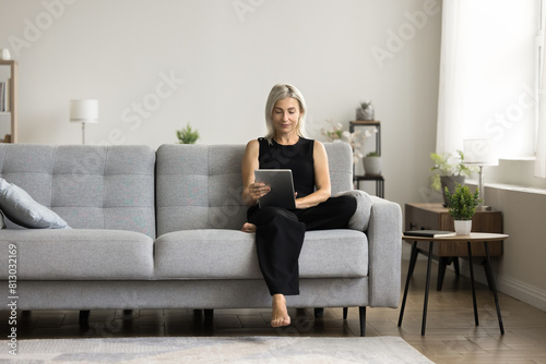 Pretty mature woman using online application on digital device, sitting with digital pad on home couch, using online application on tablet computer, reading book, enjoying domestic Internet technology