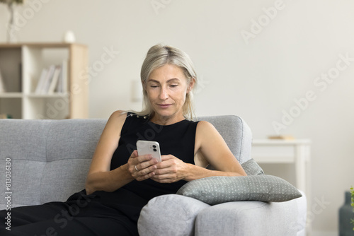 Relaxed blonde middle aged woman with cellphone resting on home couch, texting message on smartphone, chatting, typing, using online application Internet technology