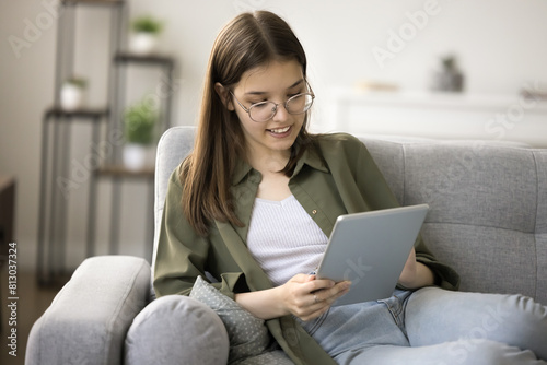 Positive smart teenager schoolkid girl in glasses using modern technology, educational app on tablet, playing online game, watching Internet content, browsing social media