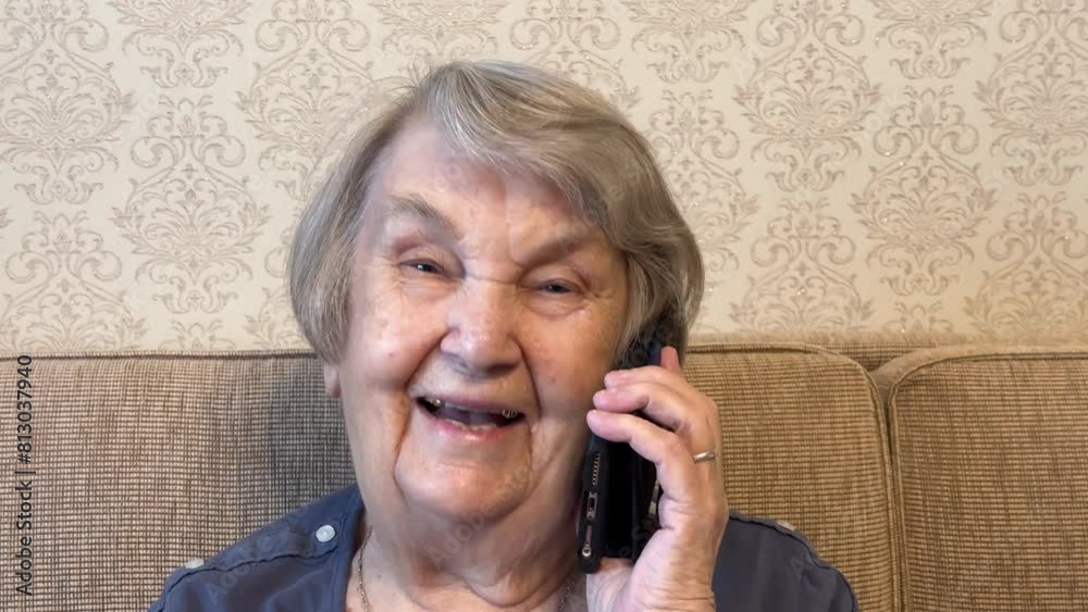 An adult retired grandmother looks into the camera and talks on her smartphone. An elderly woman 80s sitting at beige couch holding smartphone in her hand happily talking with her family. Close-up. An adult retired grandmother looks into the camera and talks on her smartphone. An elderly woman 80s sitting at beige couch holding smartphone in her hand happily talking with her family. Close-up.