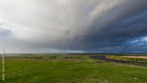  A vast landscape with a meandering river, green grassy meadow, and a horizon filled with farmland under a dramatic sky with storm clouds.