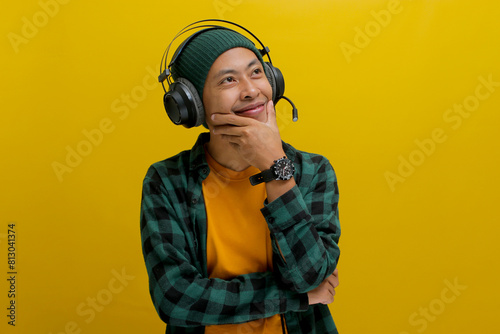 Young Asian man in a beanie and casual clothes, lost in thought while listening to music on his headphones. He gazes upward, a thoughtful smile playing on his lips. Isolated on a yellow background