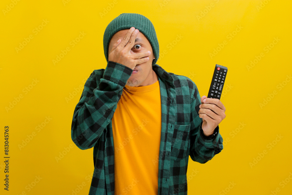 A Scared and frightened young Asian man, dressed in a beanie hat and casual shirt, holds a TV remote while watching a horror movie, peeking through his fingers, isolated on a yellow background A Scared and frightened young Asian man, dressed in a beanie hat and casual shirt, holds a TV remote while watching a horror movie, peeking through his fingers, isolated on a yellow background