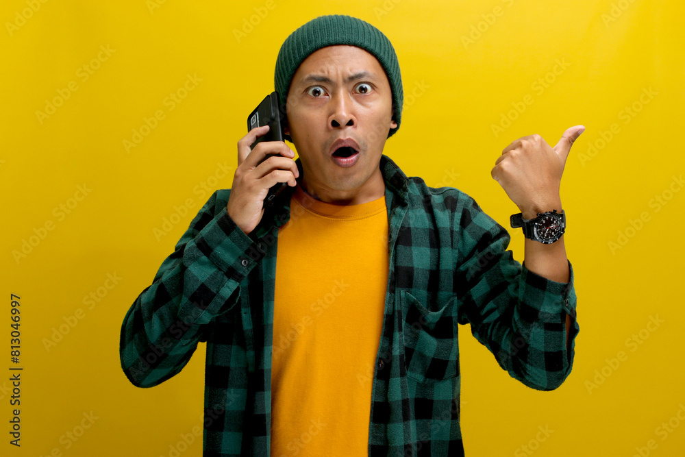 Shocked Asian man, dressed in a beanie hat and casual shirt, points to the side with his thumb at an empty space, perfect for text or advertisement, while engaged in a conversation on his mobile phone Shocked Asian man, dressed in a beanie hat and casual shirt, points to the side with his thumb at an empty space, perfect for text or advertisement, while engaged in a conversation on his mobile phone