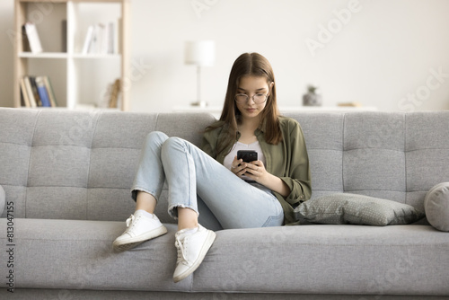 Focused gen Z teenage schoolkid girl in glasses using online educational application on smartphone, relaxing on sofa, enjoying Internet technology, leisure with digital gadget at home