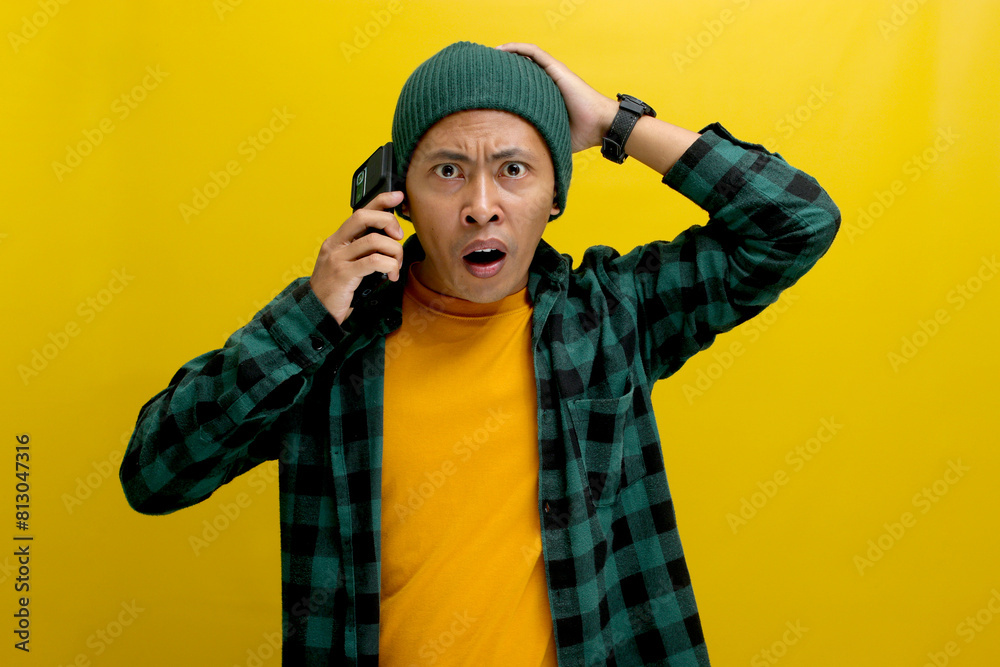 Fototapeta premium Shocked Asian man, dressed in a beanie hat and casual shirt, engages in a conversation on his mobile phone, showing a surprised expression upon receiving unexpected news, Isolated on yellow background