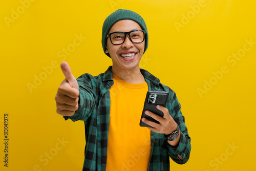 Smart young Asian man, sporting eyeglasses and dressed in a beanie hat and casual shirt, gives a thumbs up gesture while holding a phone, expressing approval, agreement, and offering a positive review photo