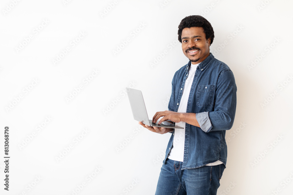 African American man is standing, holding a laptop in his hands. He is looking at the screen intently, possibly working or browsing. The background is white with copy space African American man is standing, holding a laptop in his hands. He is looking at the screen intently, possibly working or browsing. The background is white with copy space