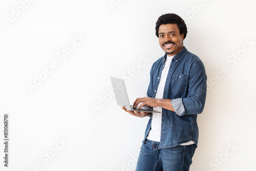 African American man is standing, holding a laptop in his hands. He is looking at the screen intently, possibly working or browsing. The background is white with copy space