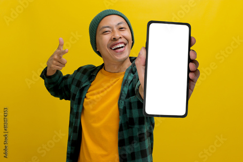 Excited Asian man, dressed in a beanie hat and casual shirt, excitedly showcases and points towards a mobile phone with an empty white screen, suggesting or recommending a new app or a special offer