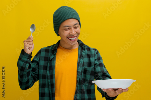 Excited young Asian man, sporting a beanie hat and casual shirt, holds a spoon while gazing at an empty plate in his hand, isolated against a yellow background