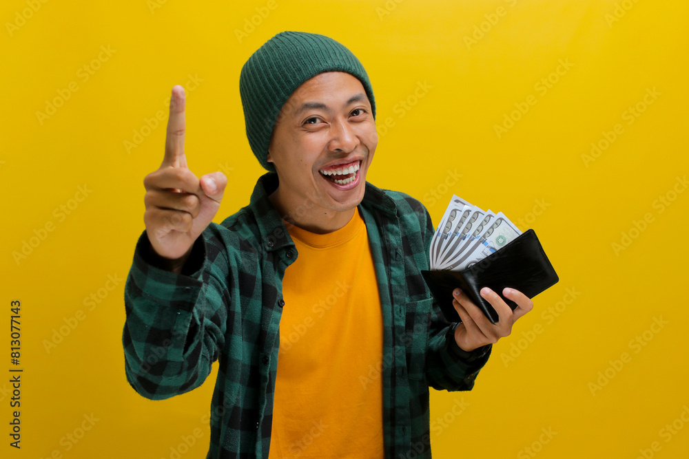 Excited young Asian man, dressed in a beanie hat and casual shirt, points his finger up, signifying a great idea or solution for saving money and making investments, while holding his wallet Excited young Asian man, dressed in a beanie hat and casual shirt, points his finger up, signifying a great idea or solution for saving money and making investments, while holding his wallet