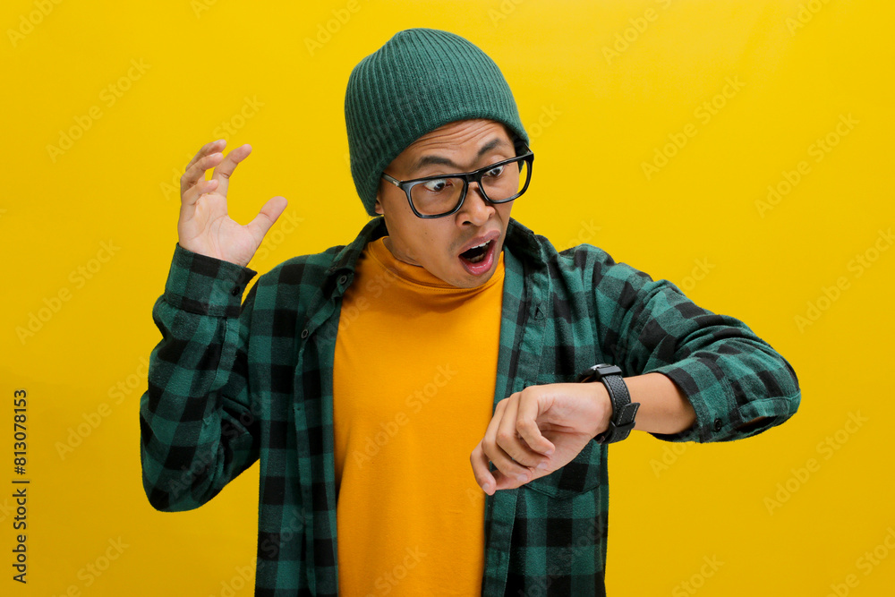 Flustered Asian man in a beanie and casual shirt checks his watch in shock. Realizing he's late, he worries about a missed appointment or deadline. Isolated on a yellow background