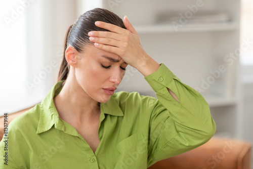 A young woman, dressed in a casual green shirt, is seated indoors with her eyes closed, evidently in discomfort as she presses her hand to her forehead, possibly indicating a headache photo