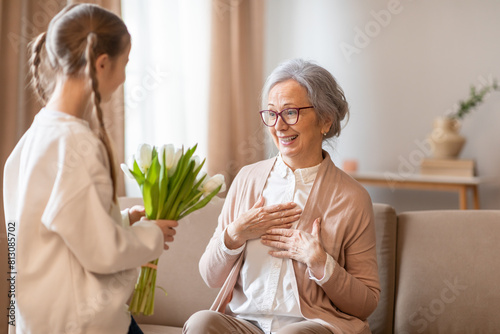 Bild auf Leinwand A young girl stands holding a fresh bouquet of tulips, extending them to an elderly woman seated on a couch