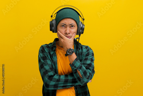 Asian man in a beanie and casual clothes, deep in thought with pensive expression while listening to music on headphones. Isolated on a yellow background. © Jamaludinyusup