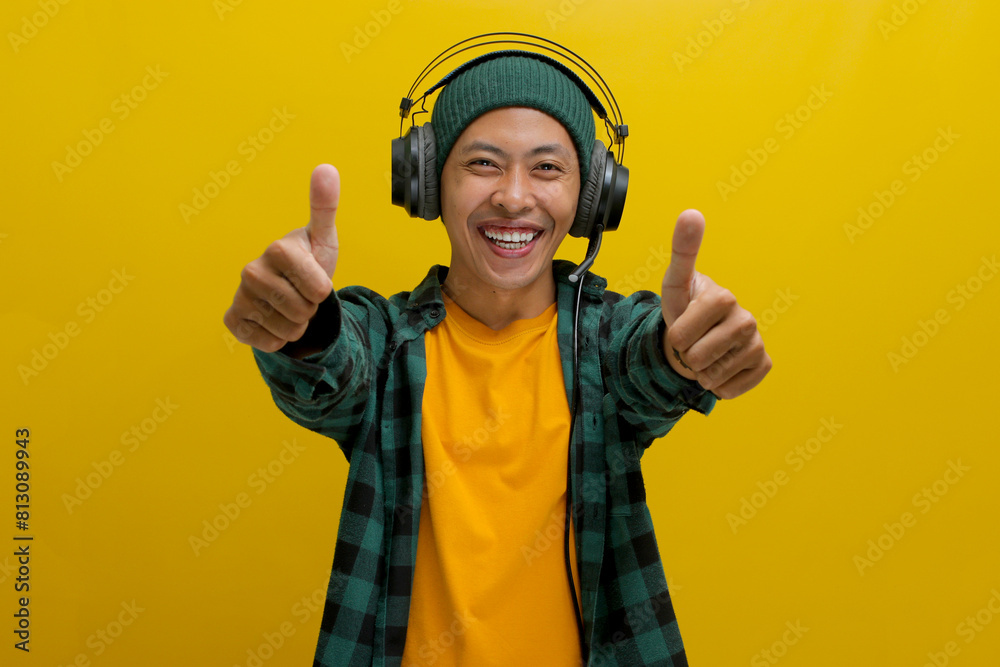 Asian man in a beanie and casual clothes smiles in approval while listening to music on his headphones. He gives a thumbs up gesture, seemingly enjoying the music. Isolated on a yellow background. Asian man in a beanie and casual clothes smiles in approval while listening to music on his headphones. He gives a thumbs up gesture, seemingly enjoying the music. Isolated on a yellow background.