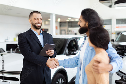 Fotografia A well-dressed salesman is shaking hands with happy Indian customers, presumably after agreeing on a sale, with a new white car in the background inside a modern car dealership showroom