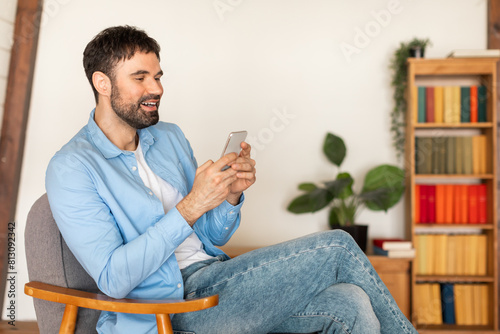 A man is seated in a chair, focused on his cell phone screen. He appears engrossed in the content displayed on the device, with his attention fully absorbed