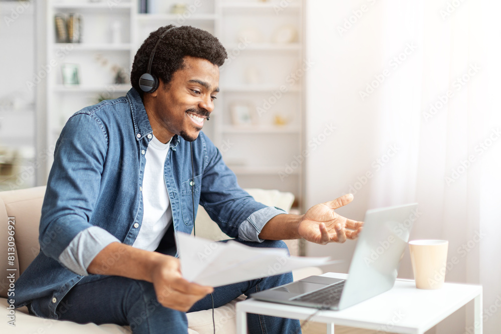 Fototapeta premium A cheerful African American man sits comfortably on a sofa, using a laptop for a virtual meeting. He is wearing headphones and holding a document