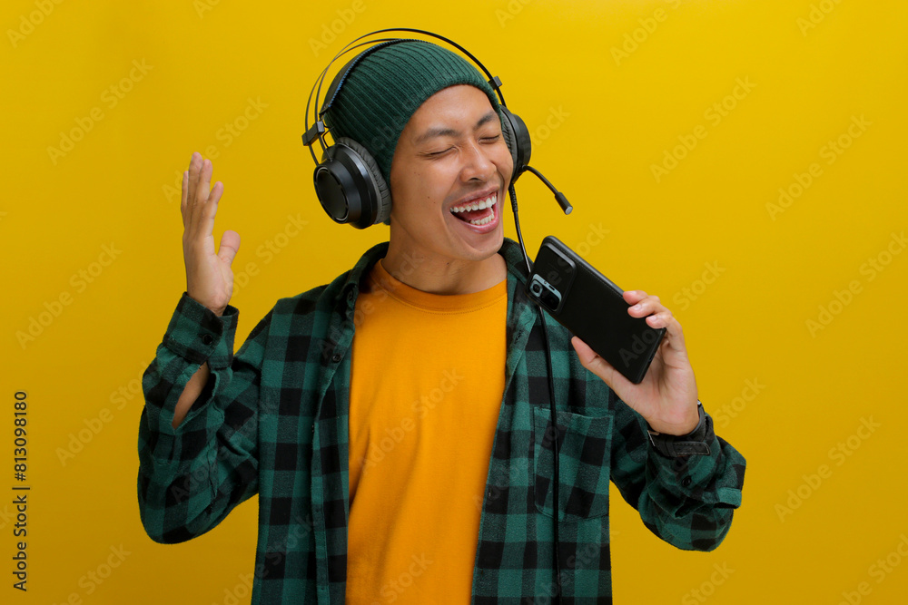 Cheerful Asian man in a beanie and casual clothes, wearing headphones and using his smartphone like a microphone. He sings along to music, enjoying a fun karaoke session at home. Yellow background. Cheerful Asian man in a beanie and casual clothes, wearing headphones and using his smartphone like a microphone. He sings along to music, enjoying a fun karaoke session at home. Yellow background.