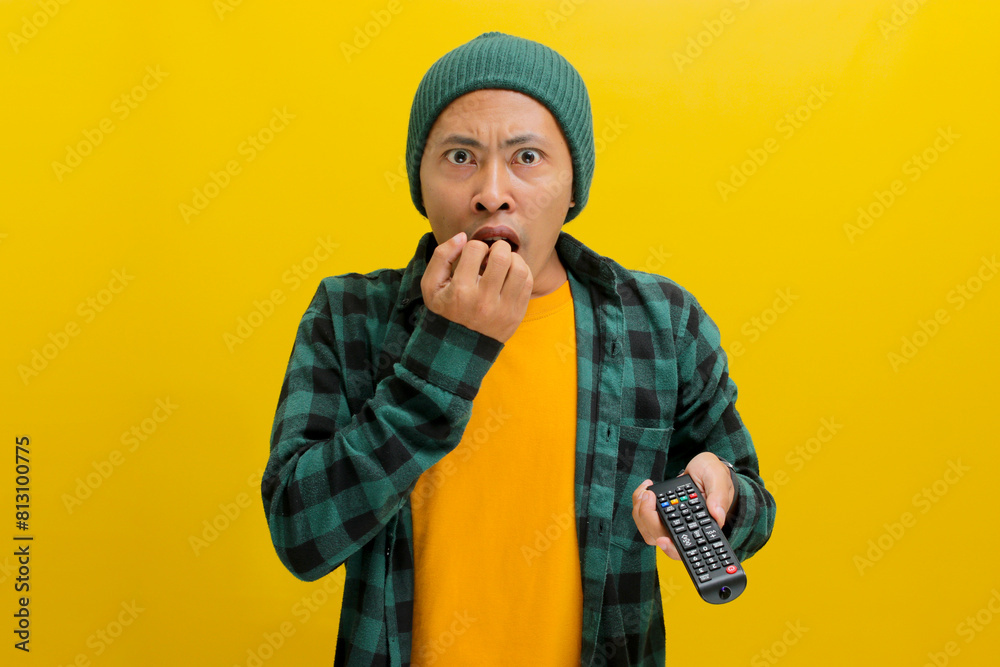 Surprised young Asian man, clad in a beanie hat and casual shirt, watches shocking news on TV while holding a remote, expressing a mix of shock, confusion, and fear, isolated on a yellow background Surprised young Asian man, clad in a beanie hat and casual shirt, watches shocking news on TV while holding a remote, expressing a mix of shock, confusion, and fear, isolated on a yellow background