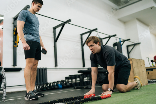 Photos Exhausted beginner sportsman falling to floor from fatigue after battle rope workout in cross training gym with help of personal trainer