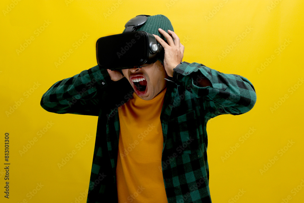 Terrified Asian man wearing a VR headset screams in fear while experiencing a horror movie in VR. Isolated on a yellow background Terrified Asian man wearing a VR headset screams in fear while experiencing a horror movie in VR. Isolated on a yellow background
