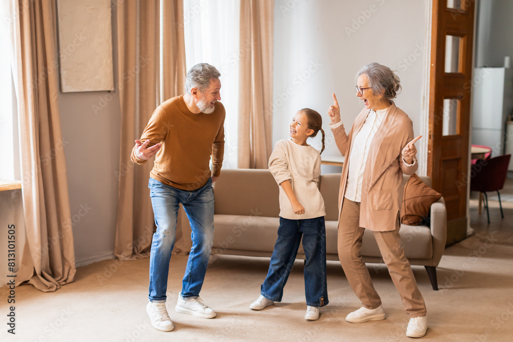 Fototapeta premium An elderly couple is joyfully dancing with a young child in the warmth of a well-lit living room. The trio appears to be having a delightful time, sharing smiles and an apparent love for music