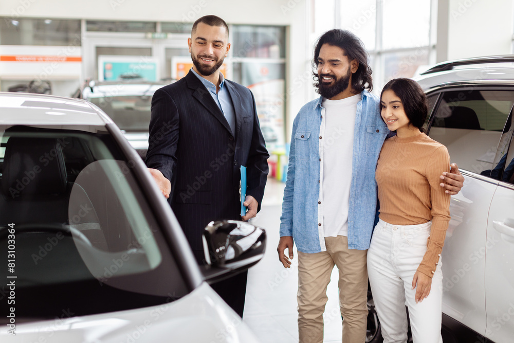 A young Indian couple is smiling and standing next to a new car at a dealership, interacting with a professional salesman who is offering the car keys A young Indian couple is smiling and standing next to a new car at a dealership, interacting with a professional salesman who is offering the car keys