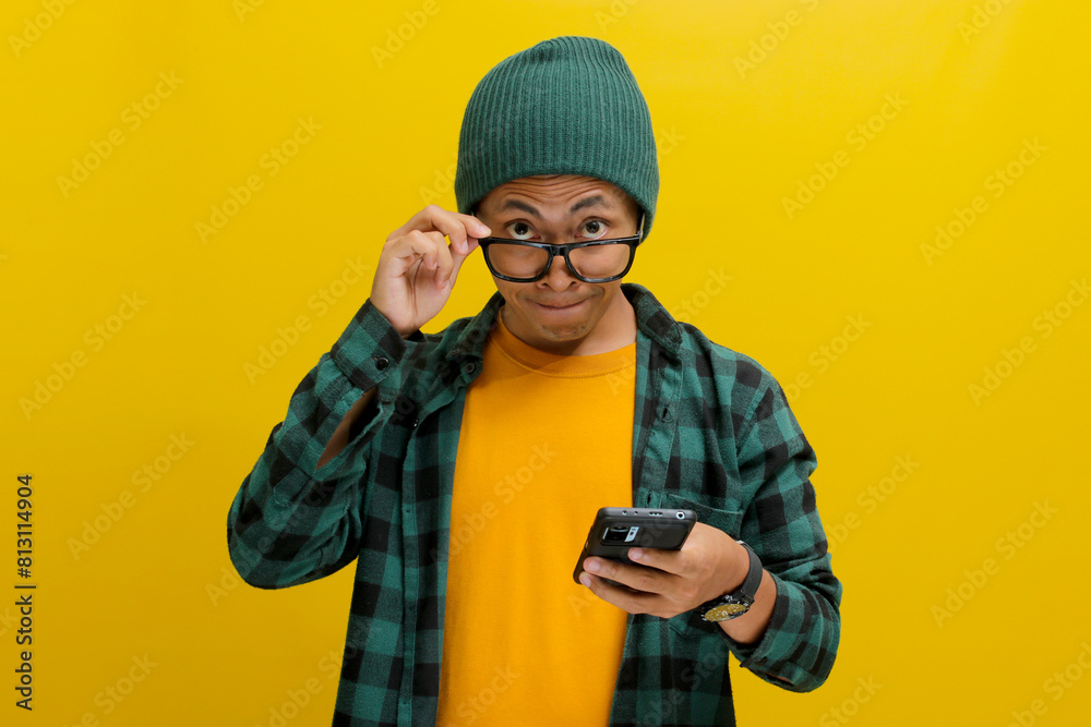 Asian man, dressed in a beanie hat and casual shirt, adjusts his eyeglasses and peeks above them, smiling as he looks at the camera with a mobile phone in hand while standing against yellow background Asian man, dressed in a beanie hat and casual shirt, adjusts his eyeglasses and peeks above them, smiling as he looks at the camera with a mobile phone in hand while standing against yellow background