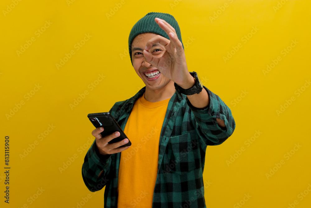 A young Asian man, dressed in a beanie hat and casual clothes, makes an OK sign gesture while holding his smartphone, conveying agreement, approval, and satisfaction, indicating a positive review