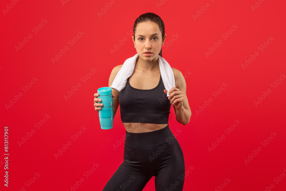 A focused, athletic woman wearing a black sports outfit and a white towel around her neck stands against a vivid red background, holding a blue water bottle, likely taking a break