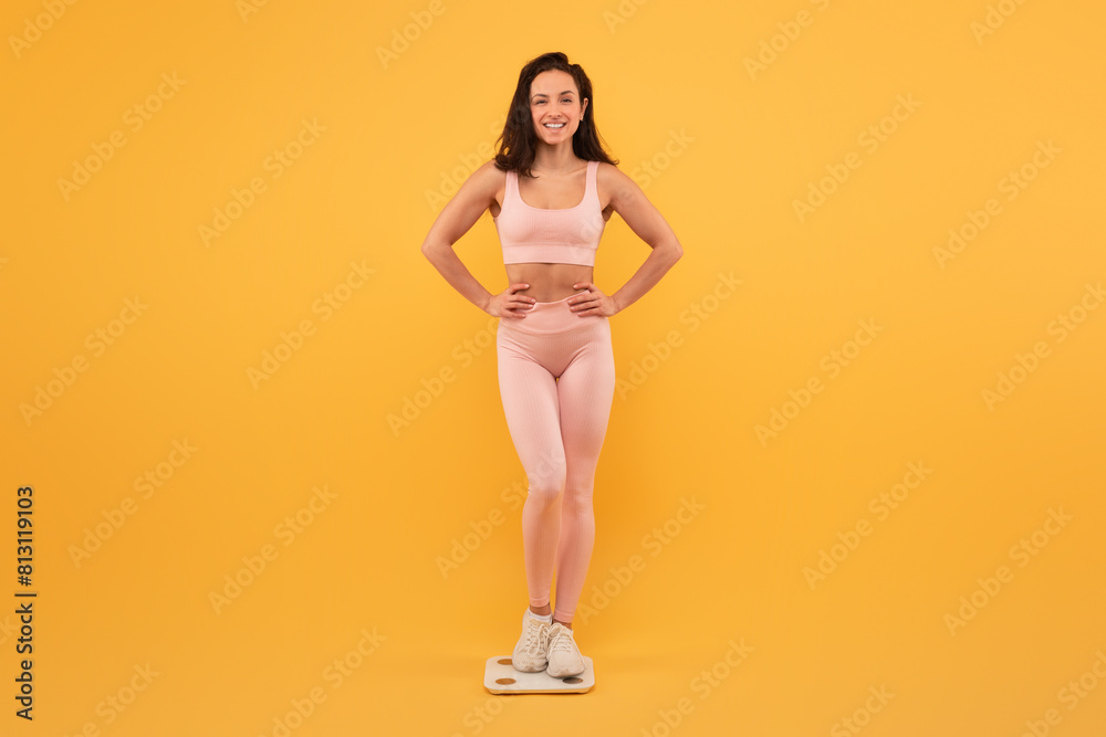 A smiling young woman stands on scale with her hands on her hips, wearing light pink activewear and white sneakers. She exudes confidence and positivity against a vibrant yellow backdrop