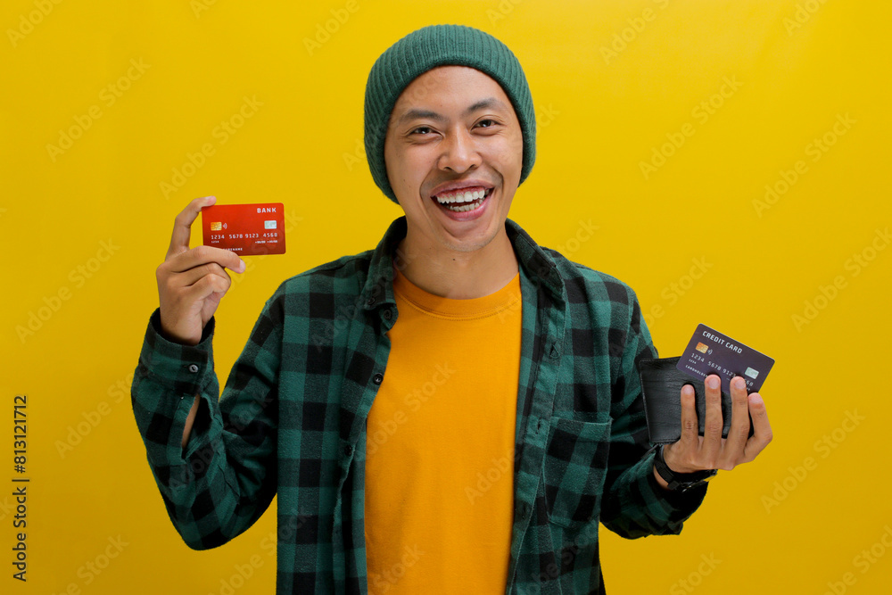 Excited young Asian man, dressed in a beanie hat and casual shirt, holds a wallet and credit card, smiling at the camera with happiness, eager to make an online payment using the credit card Excited young Asian man, dressed in a beanie hat and casual shirt, holds a wallet and credit card, smiling at the camera with happiness, eager to make an online payment using the credit card