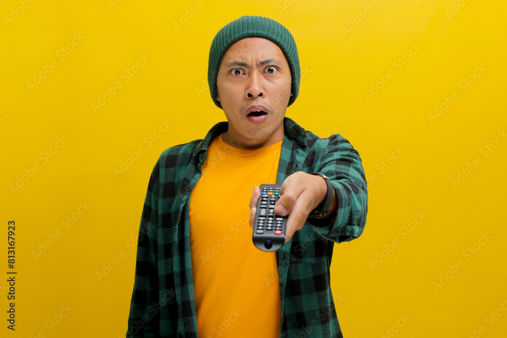 Surprised young Asian man, clad in a beanie hat and casual shirt, watches shocking news on TV while holding a remote, expressing a mix of shock, confusion, and fear, isolated on a yellow background Surprised young Asian man, clad in a beanie hat and casual shirt, watches shocking news on TV while holding a remote, expressing a mix of shock, confusion, and fear, isolated on a yellow background