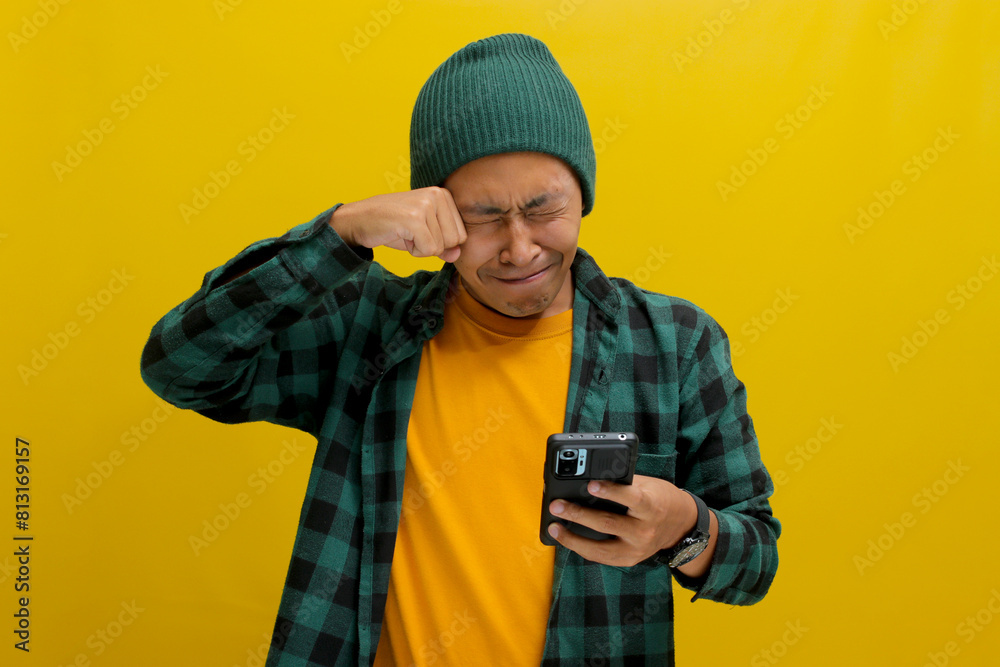 An unhappy Asian man, dressed in a beanie hat and casual shirt, holds a phone, visibly upset, displaying sadness, worry, or perhaps shedding tears in reaction to distressing news received online An unhappy Asian man, dressed in a beanie hat and casual shirt, holds a phone, visibly upset, displaying sadness, worry, or perhaps shedding tears in reaction to distressing news received online