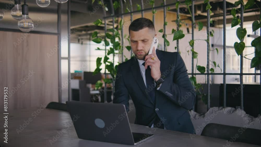 Male businessman working in a modern office at a laptop and calls on the phone. Portrait of a male boss in a formal suit. A man works intently at a laptop Male businessman working in a modern office at a laptop and calls on the phone. Portrait of a male boss in a formal suit. A man works intently at a laptop