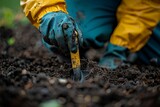 A gardener's gloved hand is pictured using a small yellow tool to work the soil in a gardening or landscaping project