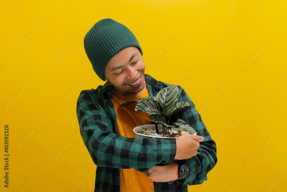 Thrilled Asian man in a beanie and casual clothes excitedly holds a Pin-stripe Calathea (Calathea ornata) houseplant in a white pot. Isolated on a yellow background. Thrilled Asian man in a beanie and casual clothes excitedly holds a Pin-stripe Calathea (Calathea ornata) houseplant in a white pot. Isolated on a yellow background.