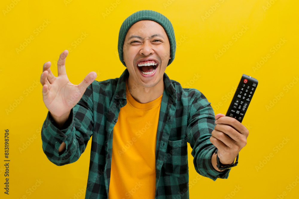 Happy Asian man, dressed in a beanie hat and casual shirt, holds a TV remote, reveling in watching a football game, passionately supporting his favorite team, while standing against yellow background